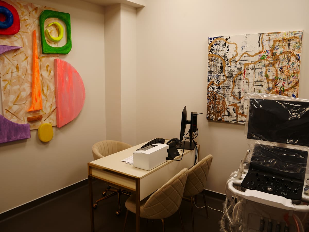 Sala de exames da AC CLINIC com equipamento de ecografia e obras de arte na parede.